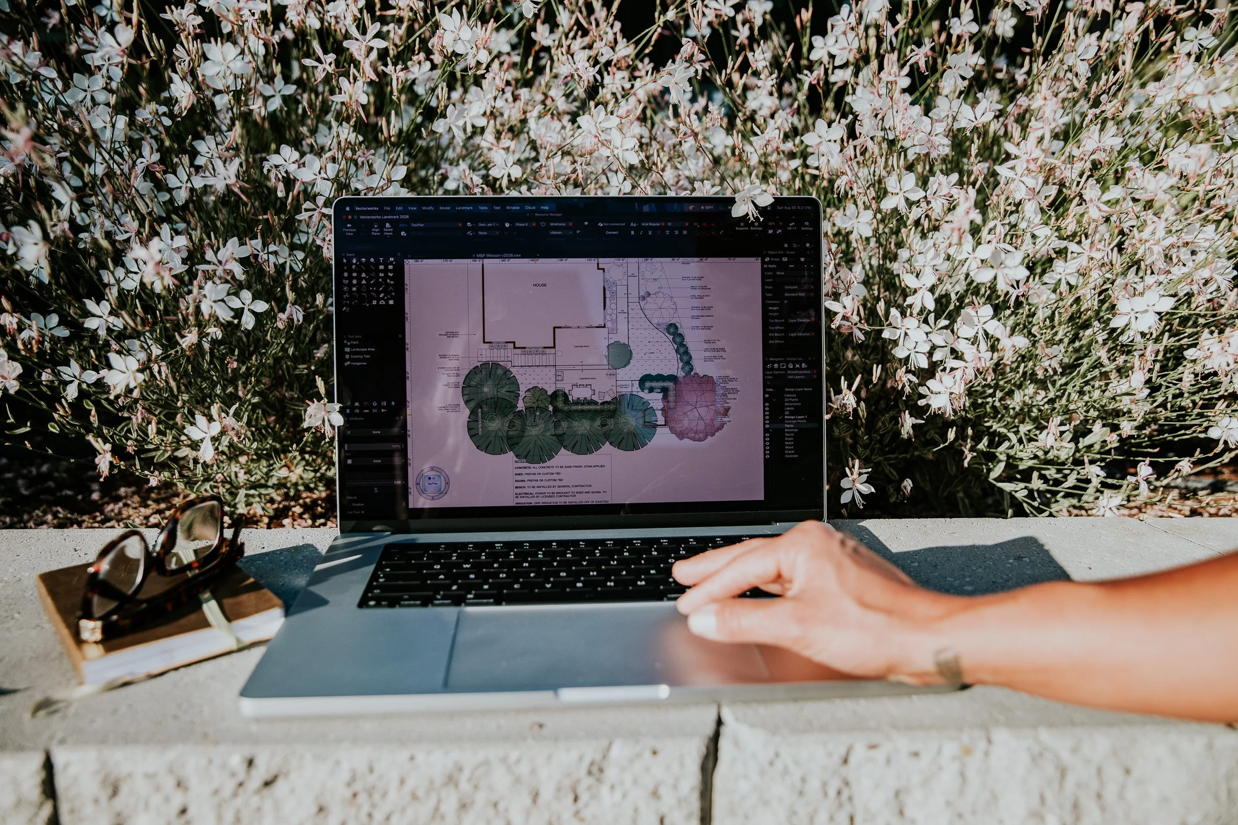 A person working on a laptop outdoors surrounded by blooming white flowers, with sunglasses and a notebook nearby.