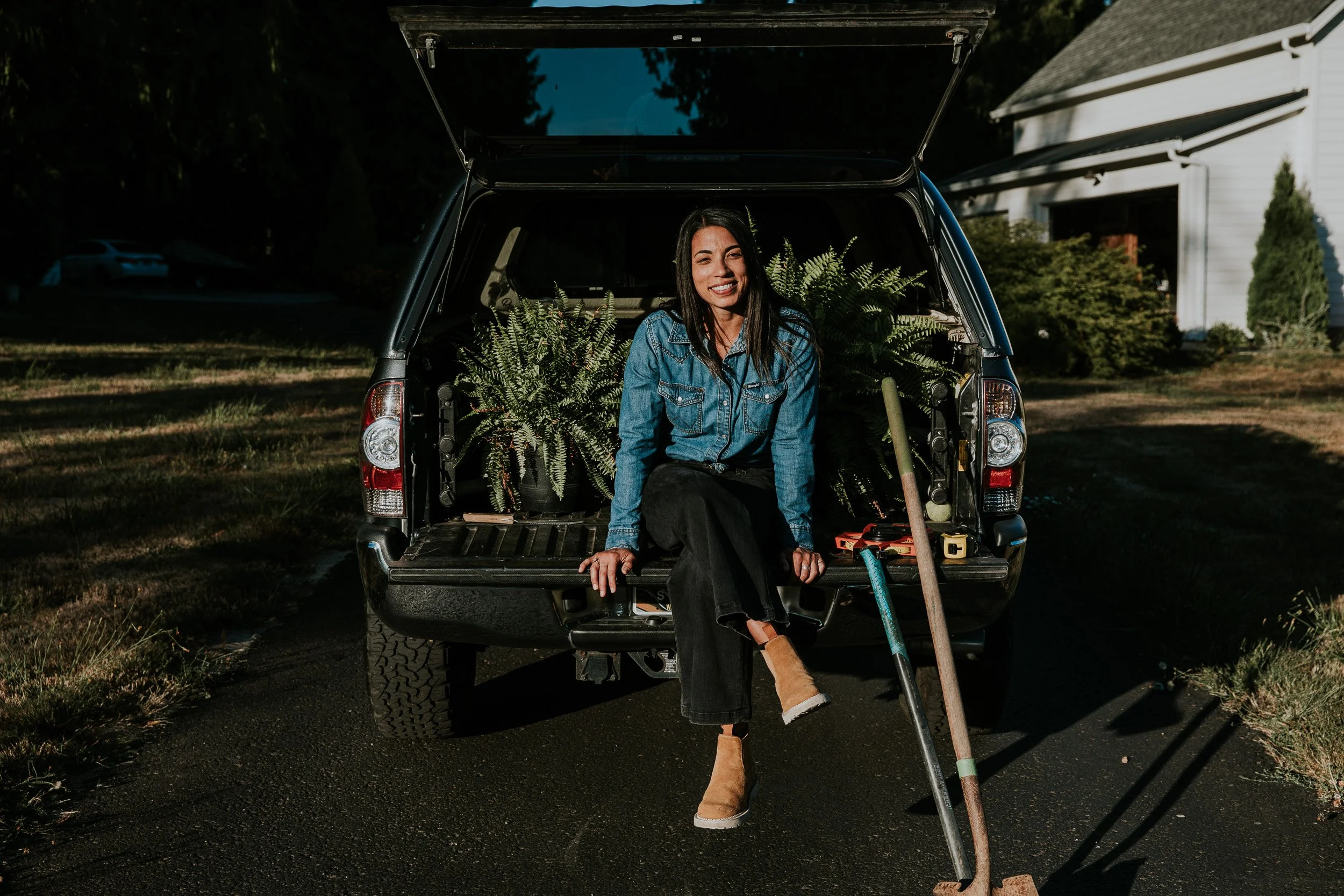 A woman sitting on the open tailgate of a black vehicle with plants behind her, smiling, wearing a denim jacket and black pants, with gardening tools and a potted plant nearby, in a suburban yard during daylight.