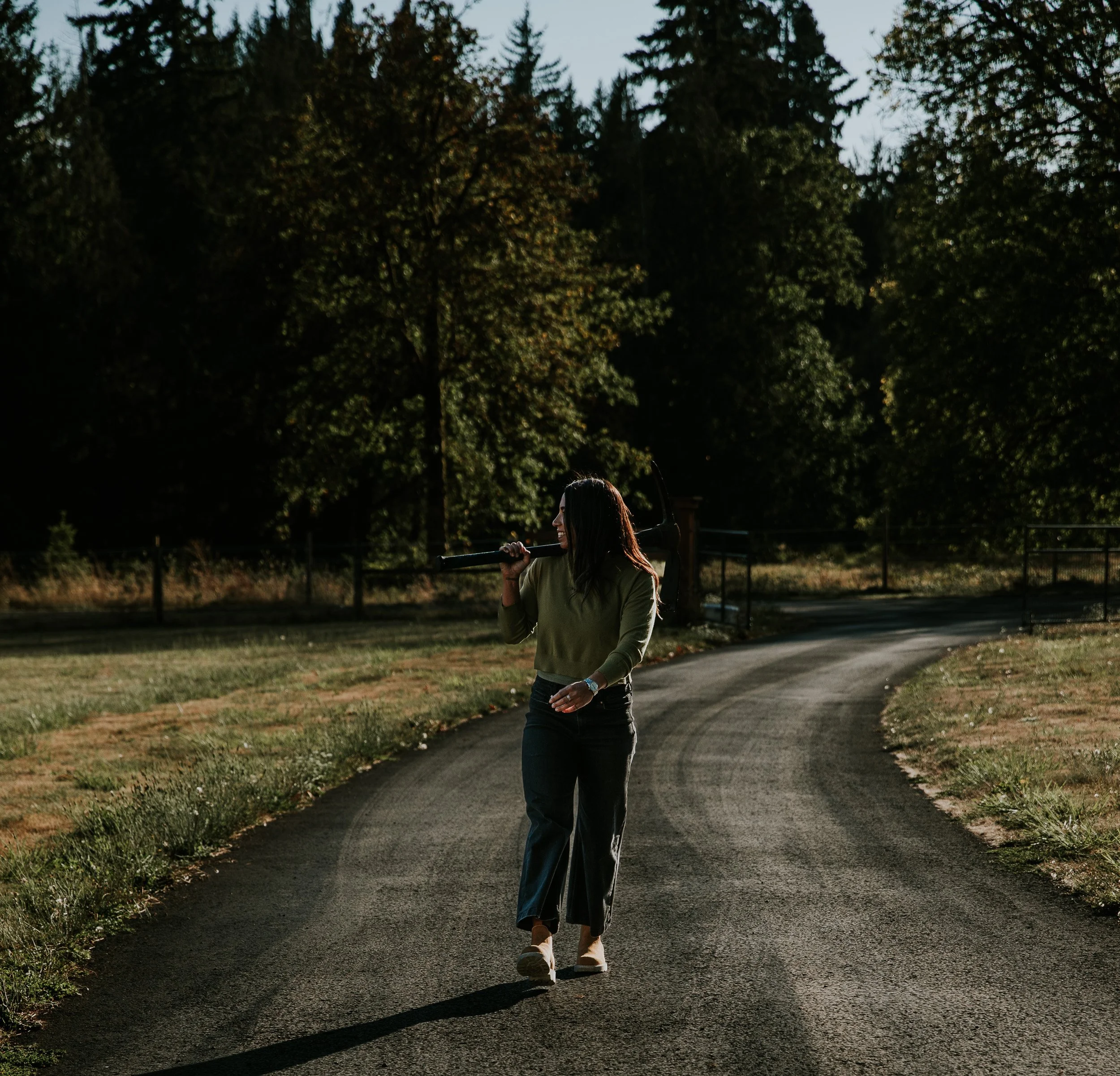 A woman with long dark hair walks along a paved path with a baseball bat over her shoulder in a park during late afternoon or early evening, with tall trees and a fence in the background.