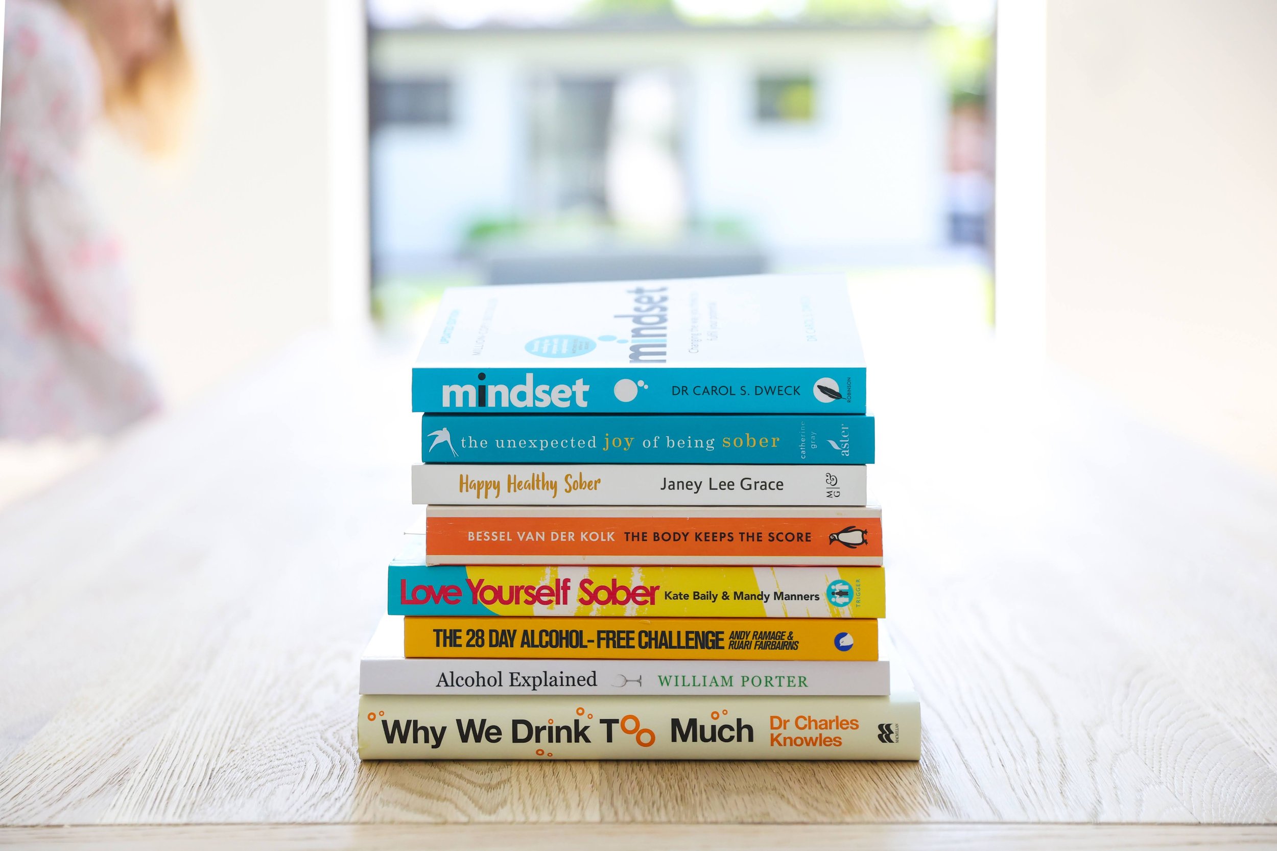 A stack of books about sobriety and addiction on a wooden table, with a blurred background of a bright room with large windows.