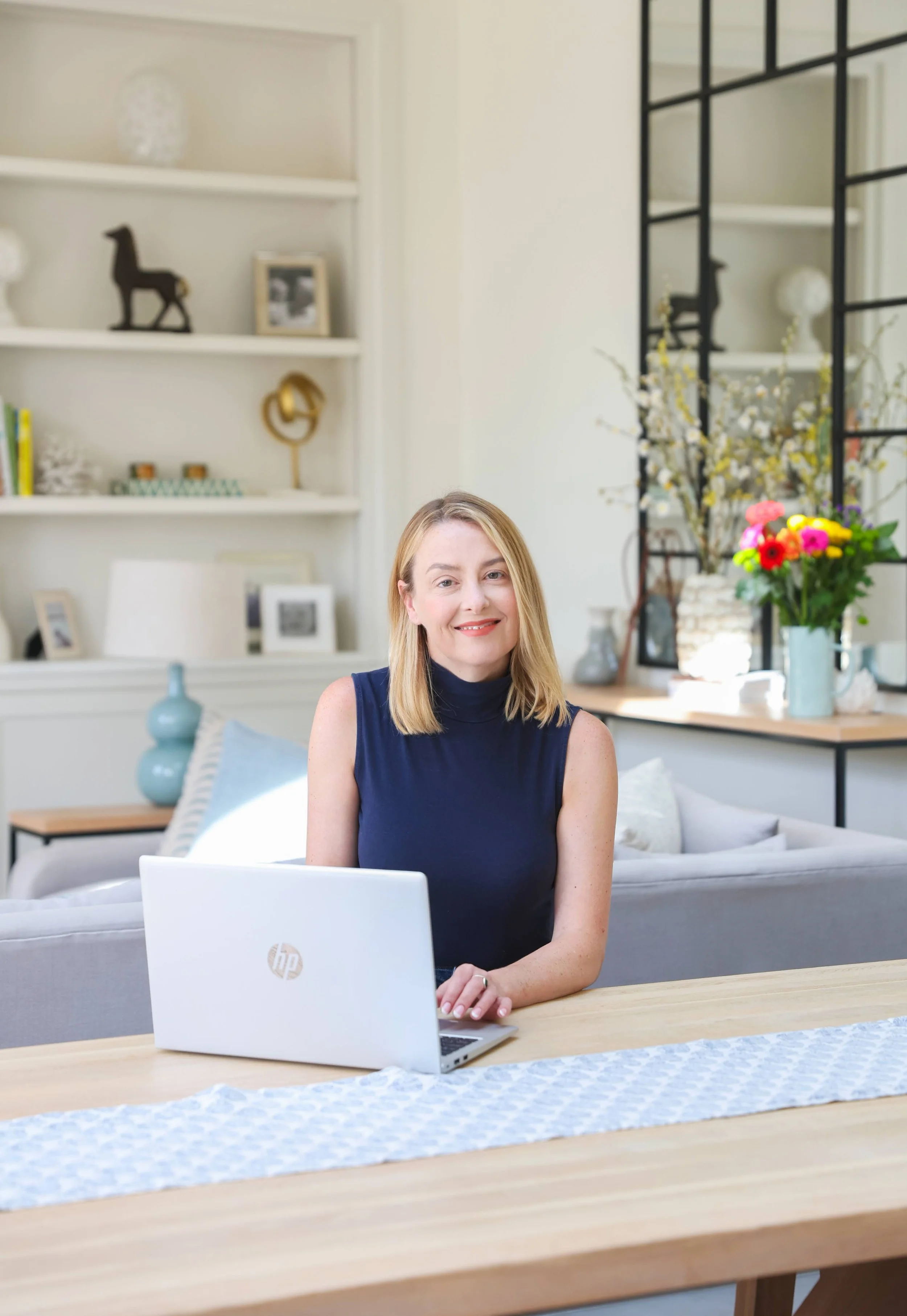 A woman sitting at a wooden dining table with an open laptop in front of her in a bright, modern living room with decorative shelves, framed photos, and fresh flowers.