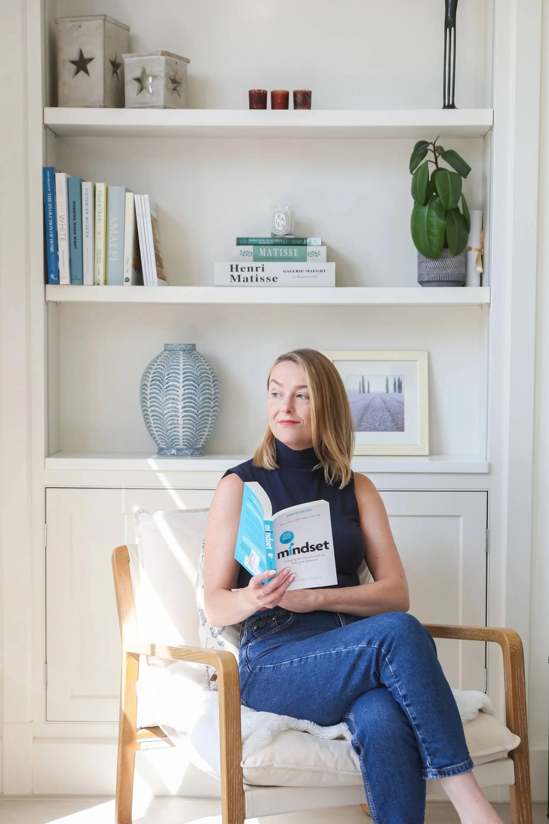A woman sitting on a chair, reading a book titled 'mindset', in a room with a white bookshelf, decorative items, and a framed picture.