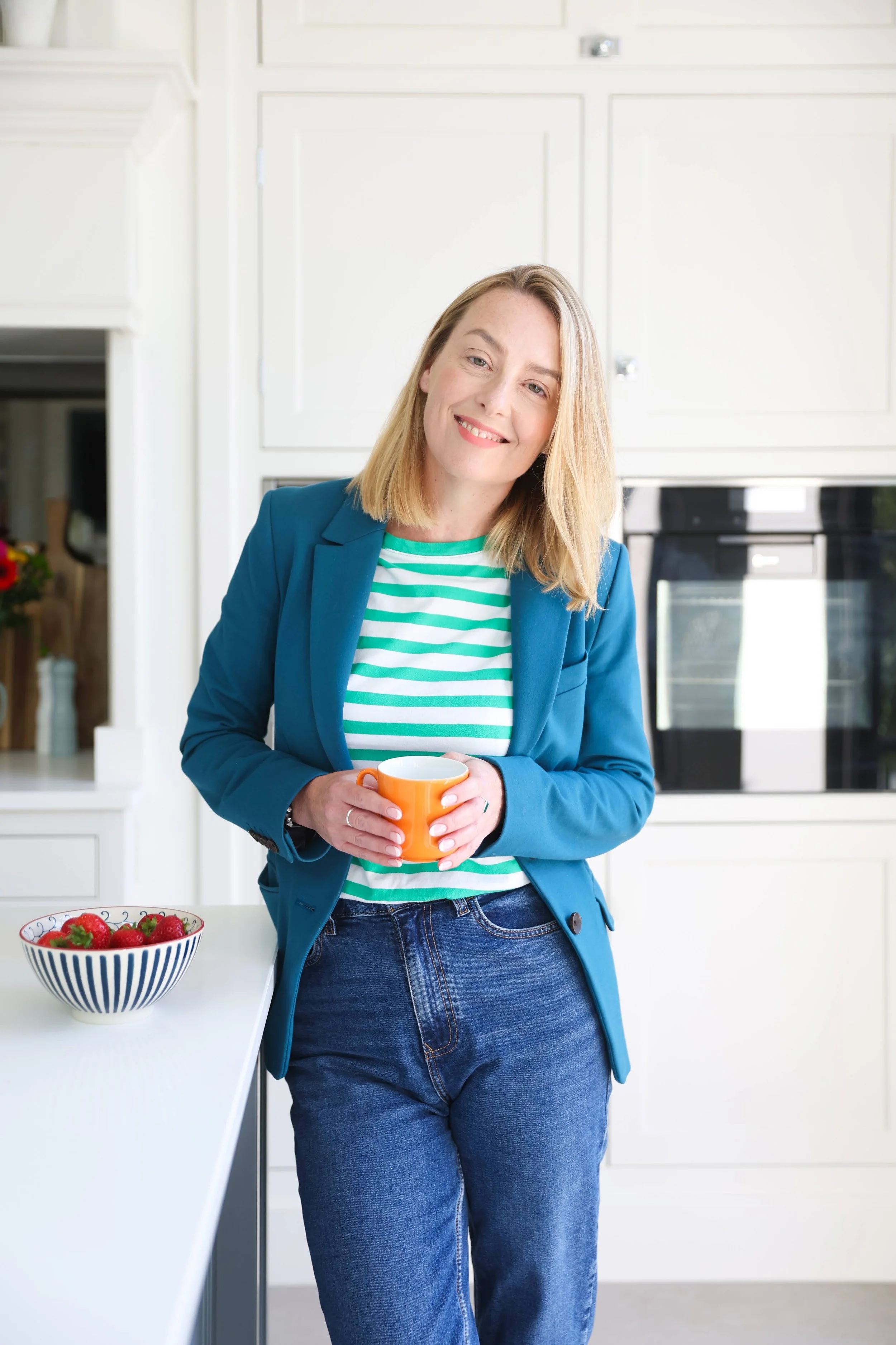 A woman with blonde hair wearing a blue blazer and striped green and white shirt standing in a bright kitchen, holding an orange mug and smiling, with a bowl of strawberries on the white kitchen counter.
