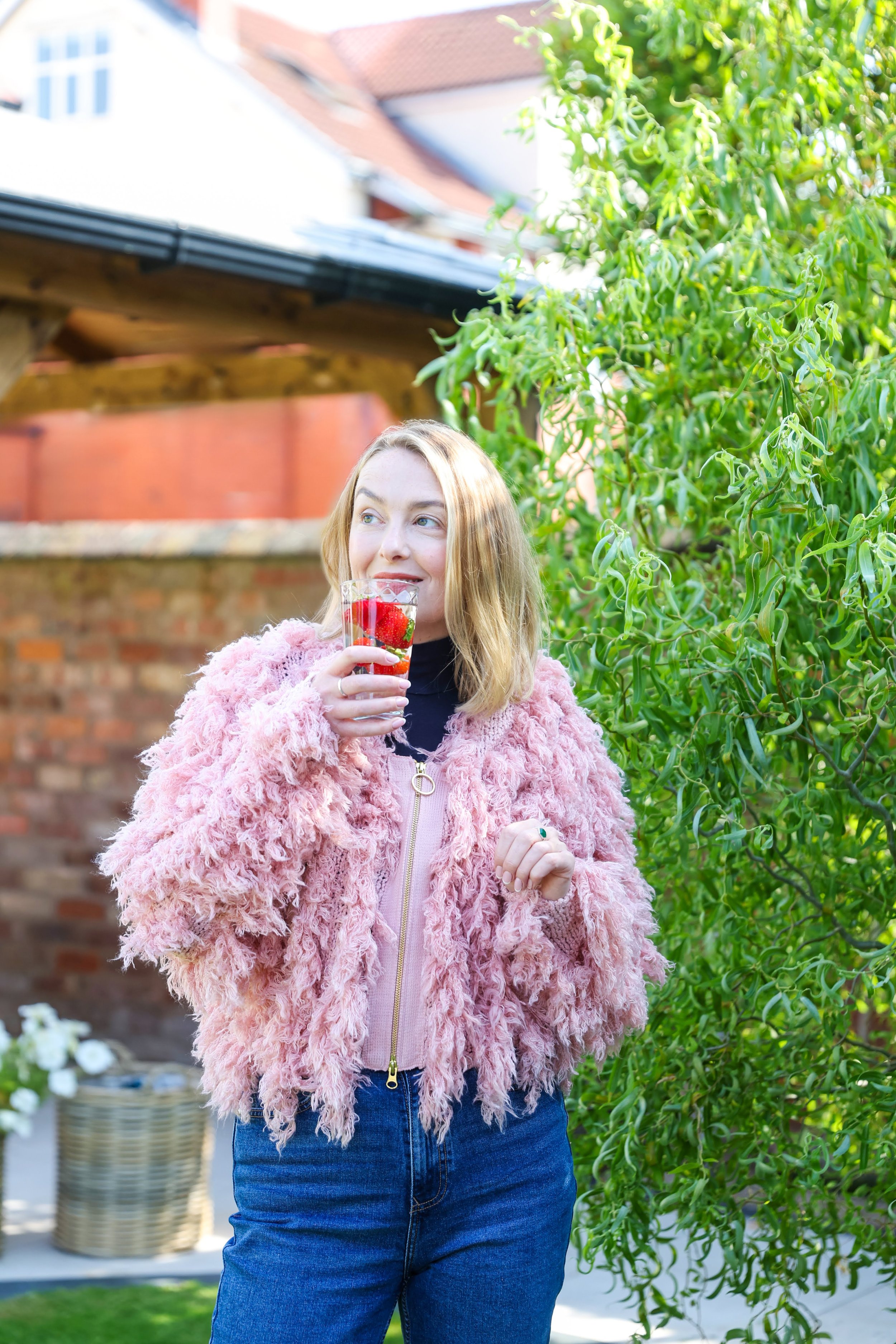 A woman with blonde hair wearing a pink fuzzy jacket and blue jeans, holding a glass of strawberry-infused drink, standing outdoors in a garden.