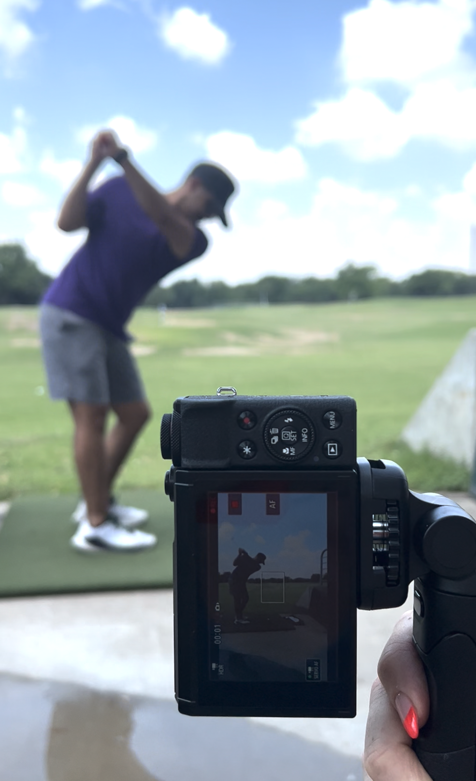 A person taking a photo of a golfer on a golf course, with the camera screen showing the golfer in a ready stance preparing to swing.