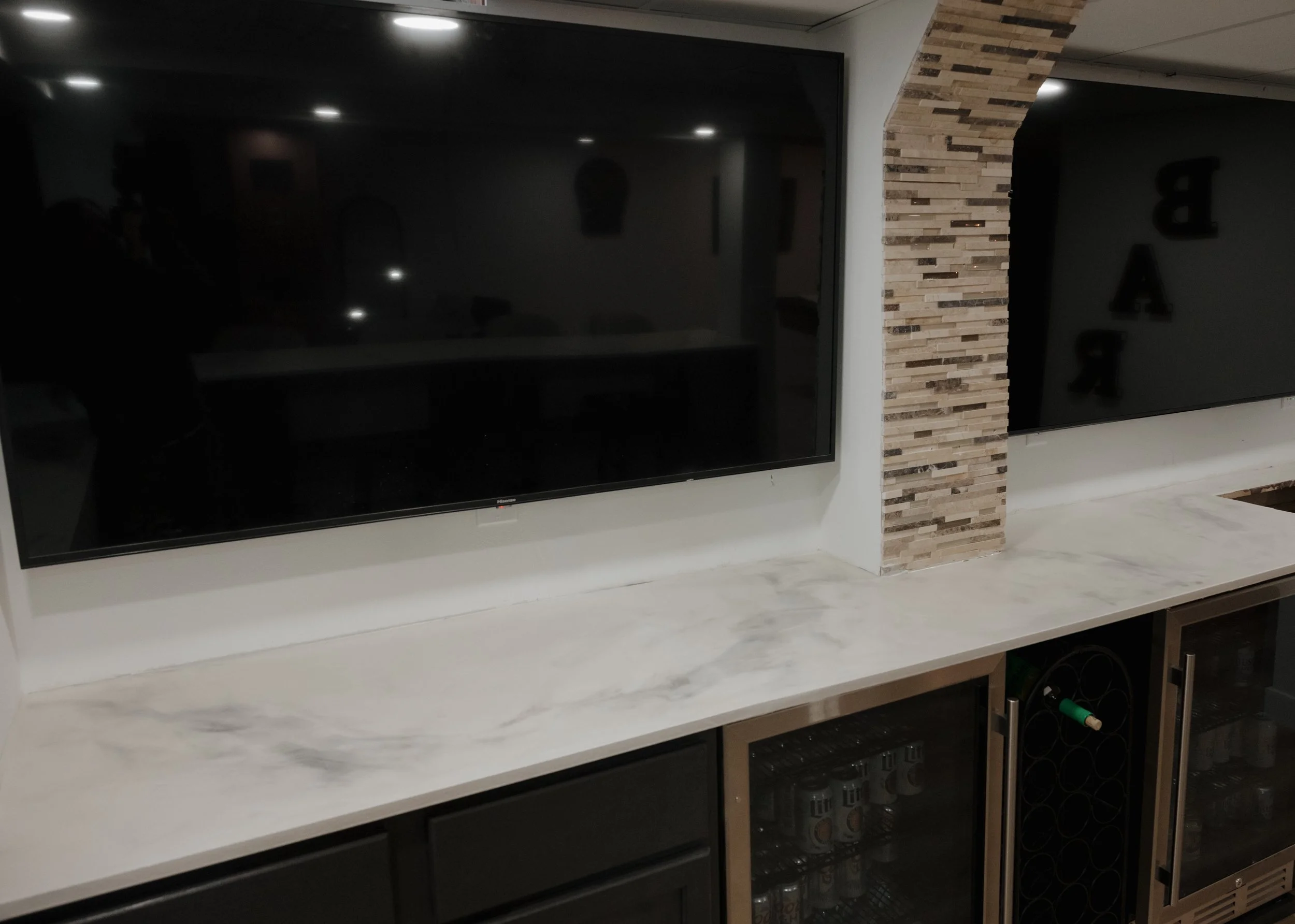 Living room with two large blank black wall-mounted screens, one looks like a television, and a textured brick wall division. There is a white marble or quartz countertop below the screens, with a wine cooler and some bottles stored underneath.