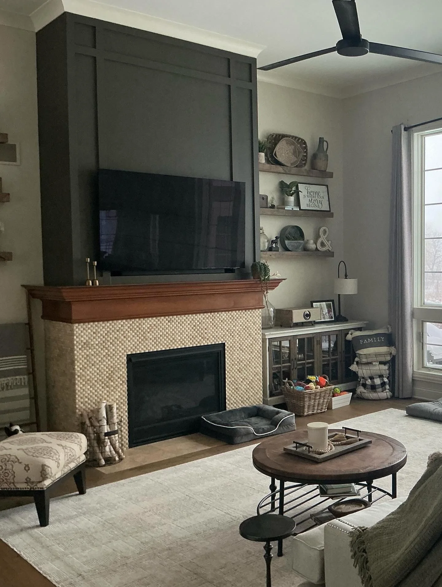 Living room with fireplace, wall-mounted TV, sofa, patterned armchair, coffee table, basket of toys, shelves with decorative items, large window with curtains, ceiling fan, and neutral-colored walls and flooring.