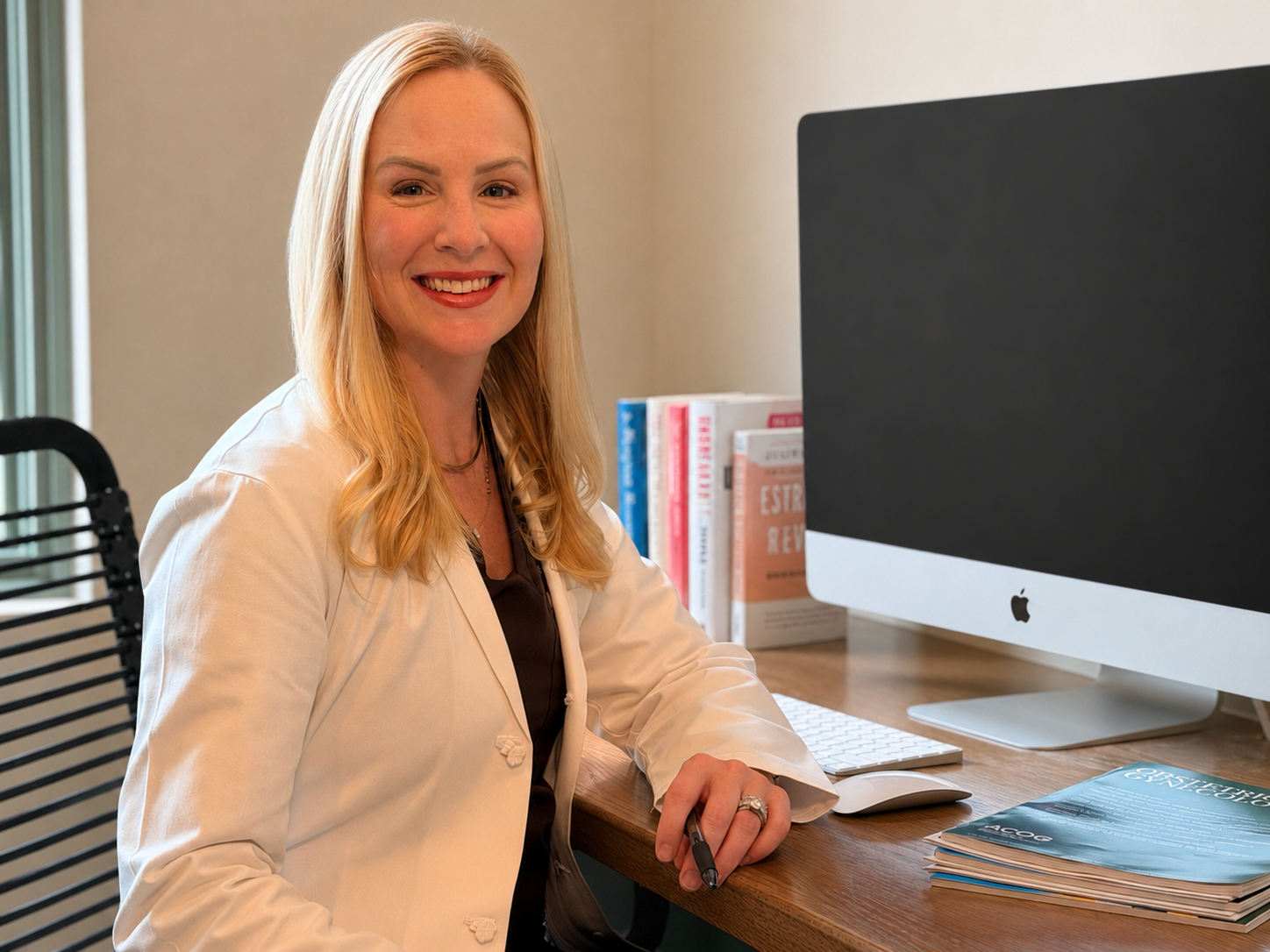 A woman with long blonde hair, wearing a light-colored blazer, sitting at a wooden desk with an Apple computer, books, and magazines, smiling at the camera.
