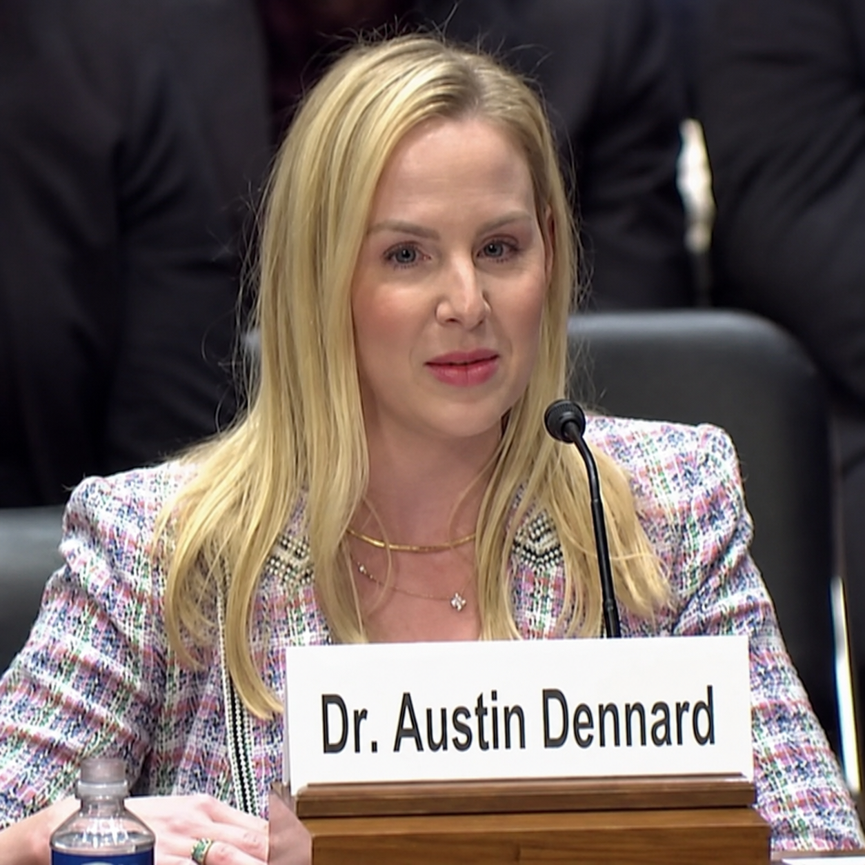 Woman with blonde hair speaking into a microphone at a hearing, wearing a colorful jacket and a gold necklace. There is a nameplate in front of her that reads 'Dr. Austin Dennard'.