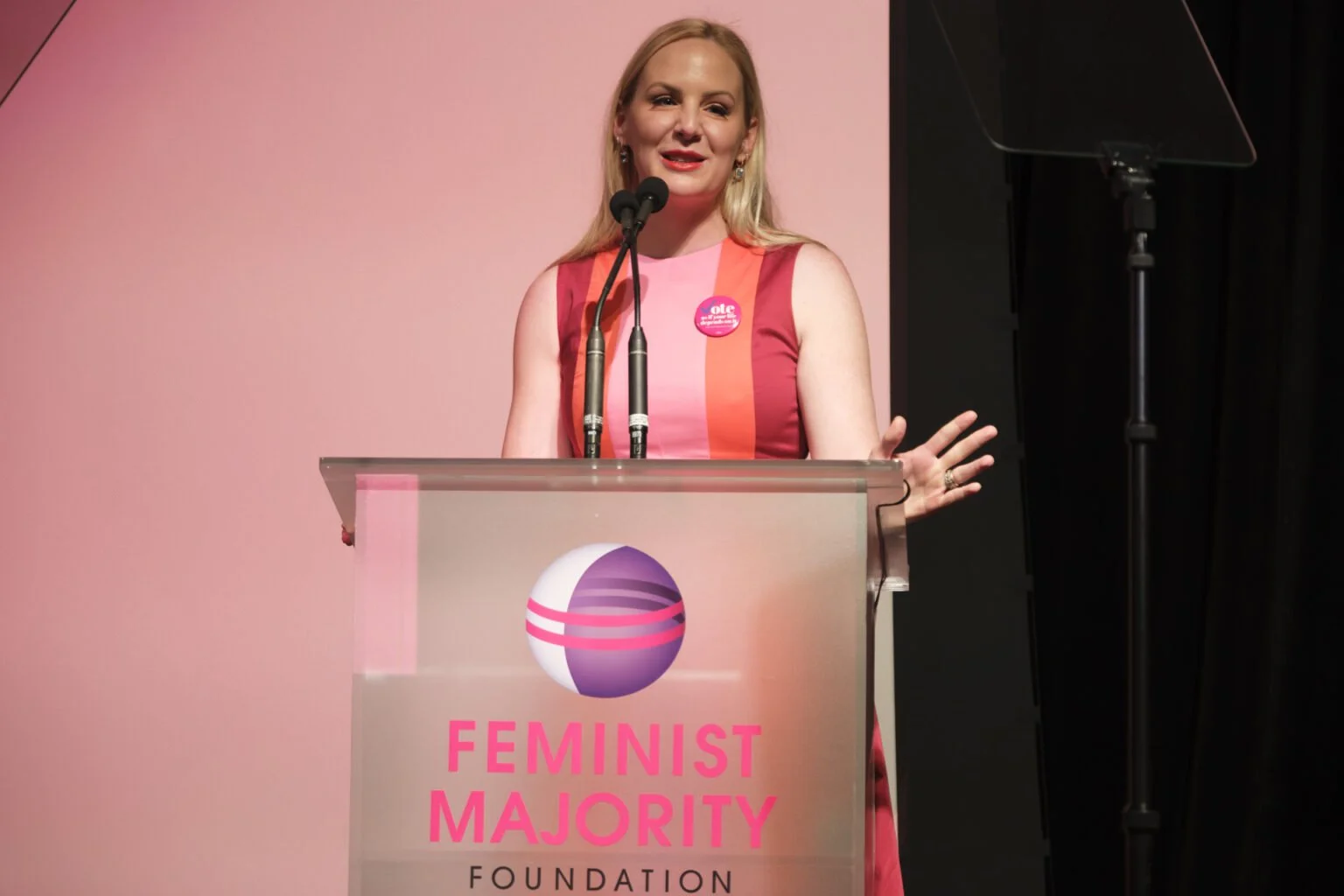 A woman speaking at a podium with a sign reading "Feminist Majority Foundation" and a logo featuring a purple, pink, and white sphere. She is wearing a sleeveless pink and red dress and a pink button.