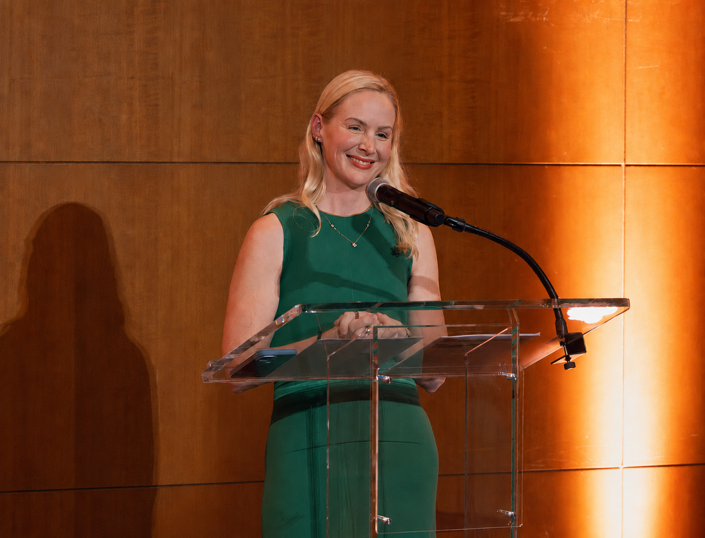 A woman with blonde hair wearing a green dress and a necklace standing at a clear podium with a microphone, smiling while speaking in front of a wooden wall background.