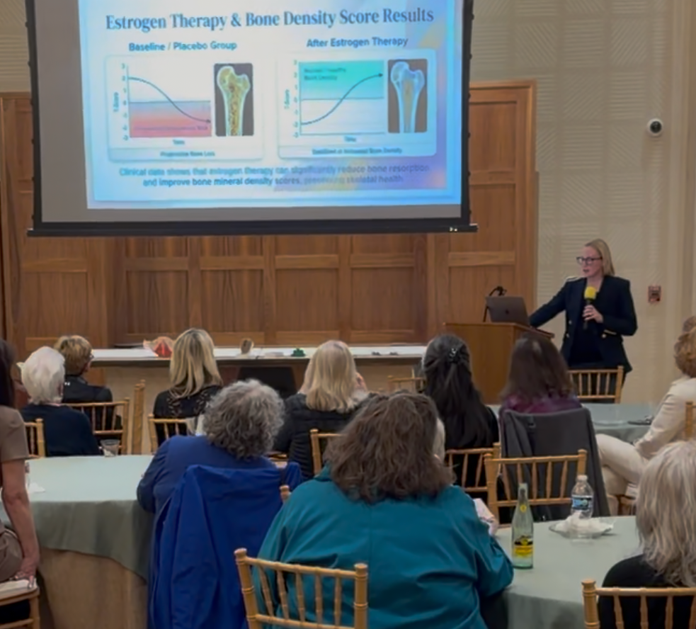 A woman in a black blazer and glasses presents a slide about estrogen therapy and bone density scores to an audience at a conference or seminar. The slide shows two graphs comparing bone density before and after estrogen therapy with images of bones.