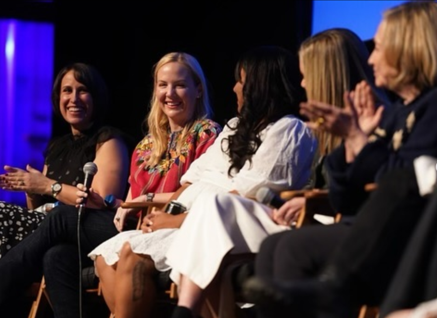 A group of six women sitting in a panel or audience, smiling and clapping, during an event or conference.