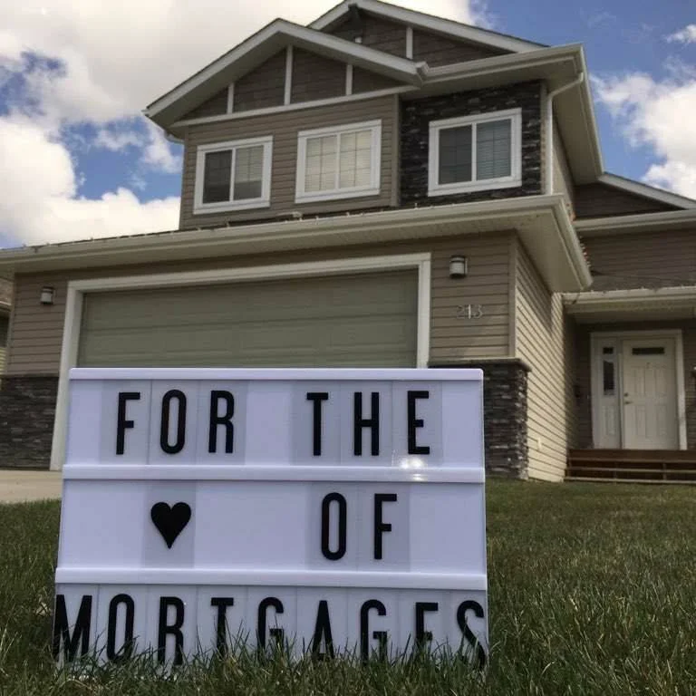 A house with a light-colored exterior, multiple windows, and a green garage door. A sign in the front yard reads, "For the Love of Mortgages," with a heart symbol. Mortgage specialist. Mortgage expert. Central Alberta.