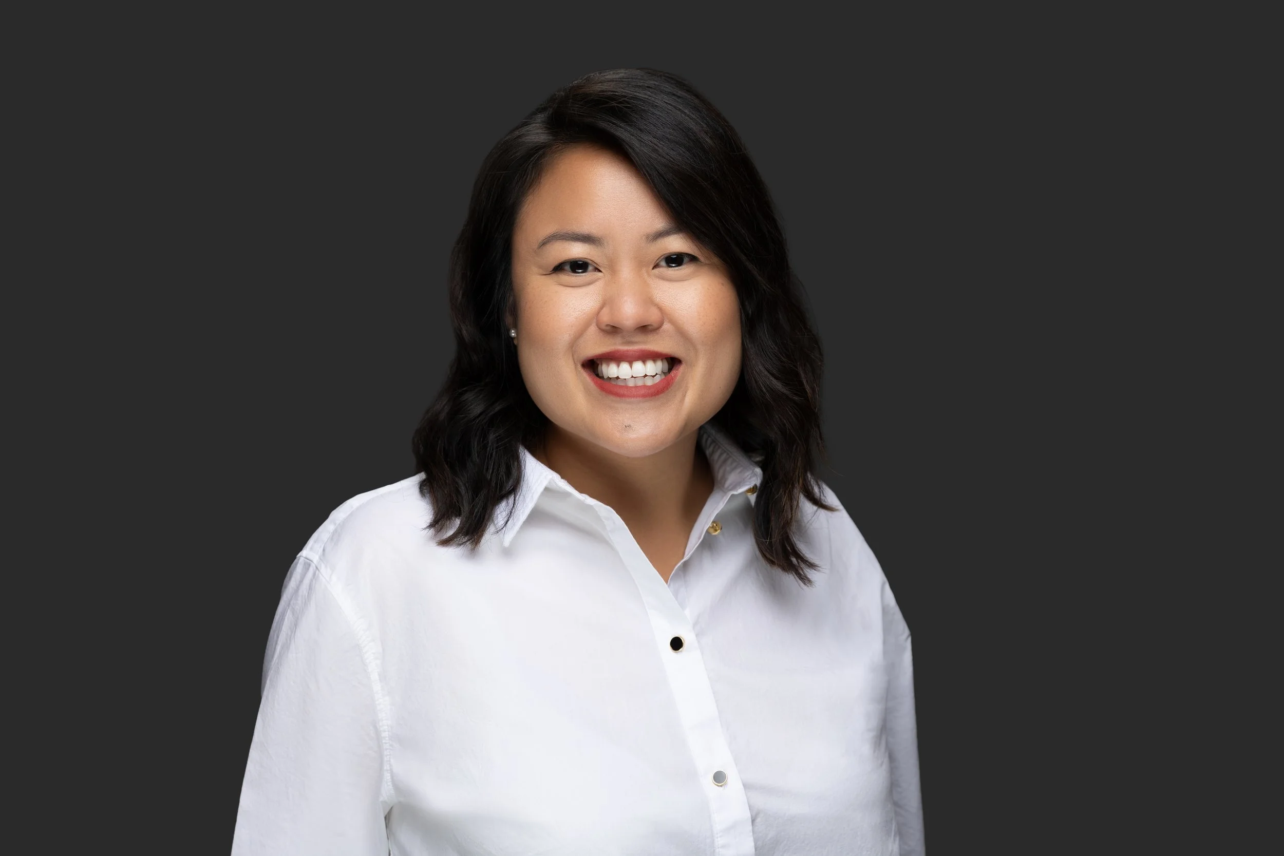 Headshot of a smiling woman with medium-length dark hair wearing a white button-up shirt, against a gray background.