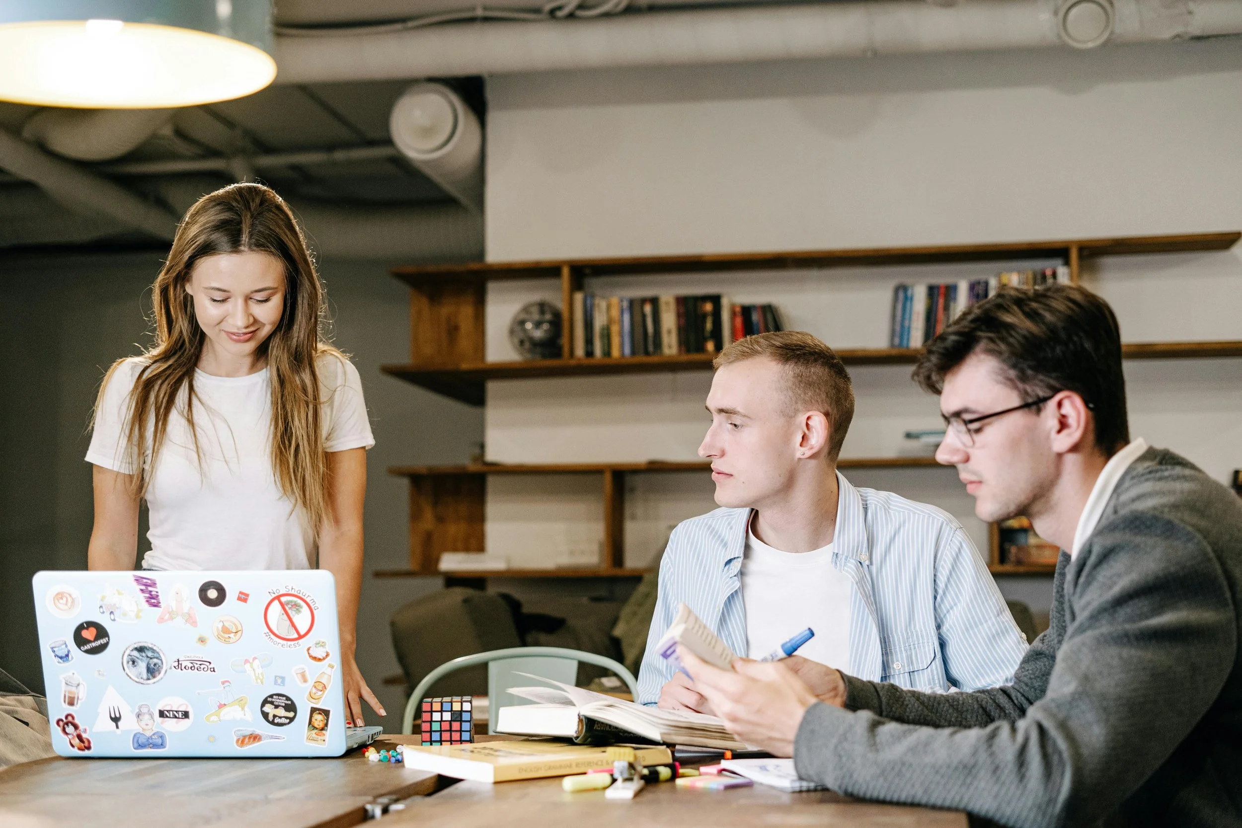 Four young adults sitting at a table working together, with one standing. The table has books, notebooks, a Rubik's cube, and a laptop covered in stickers.