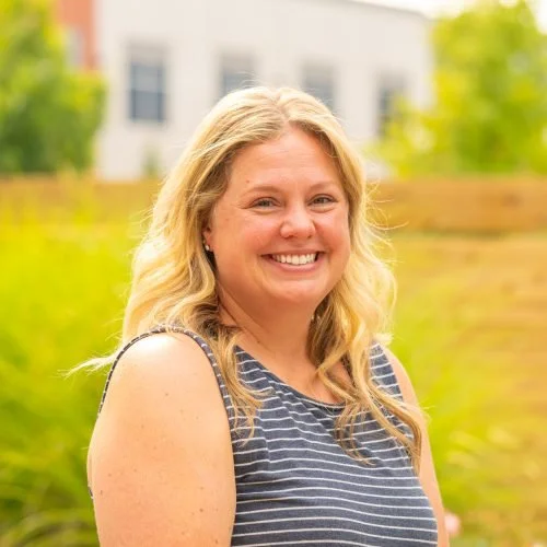 A smiling woman with blonde hair wearing a sleeveless striped top standing outdoors with a blurred background of greenery and a building.