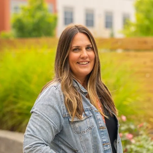 A woman with long brown hair wearing a denim jacket outdoors with blurred greenery and buildings in the background.