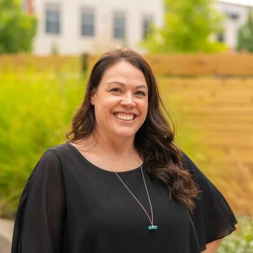 Smiling woman with long dark hair wearing a black shirt and necklace outdoors with a house and green trees in the background.