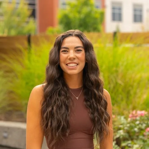 Young woman with long wavy dark hair smiling outdoors in a garden with green plants and a building in the background.