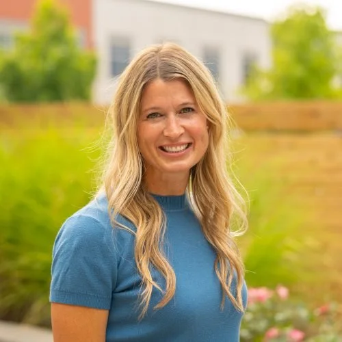 A woman with long blonde hair smiling outdoors, wearing a blue shirt with a blurred background of greenery and a building.
