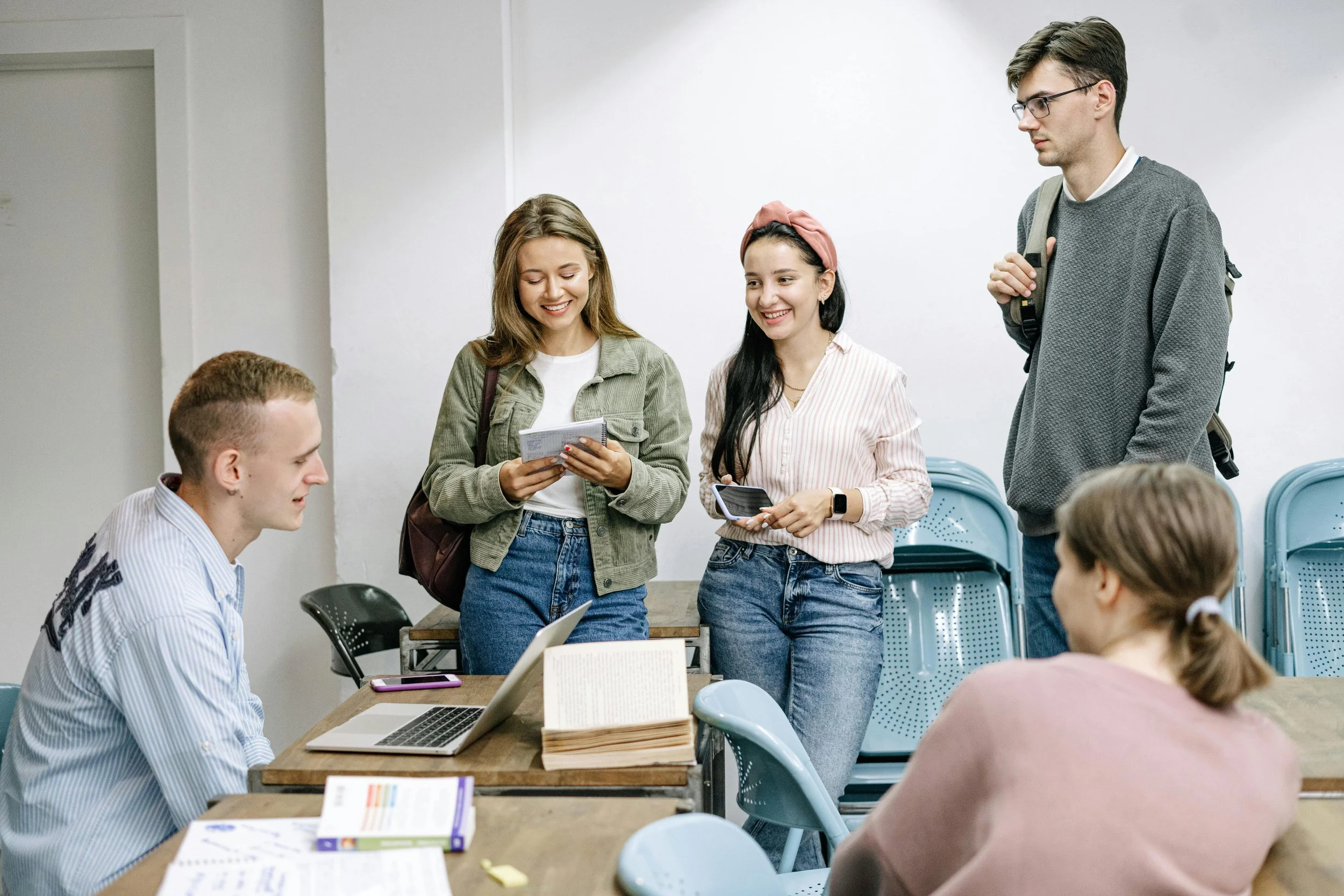 Group of students in a classroom, with two girls smiling and talking, and boys seated at desks with laptops and books.