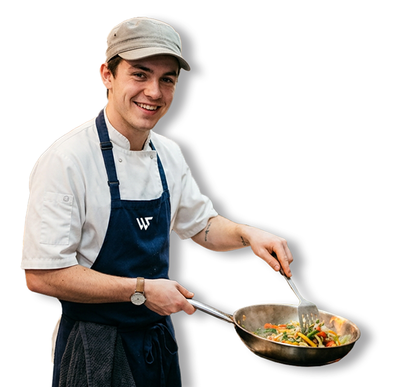 Young male chef cooking with a skillet, smiling, wearing a beige cap, white chef coat, blue apron, and a watch.