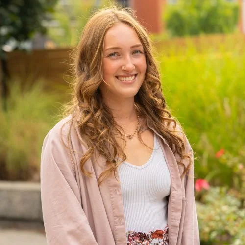 A young woman with long, curly blonde hair smiling outdoors in a garden or park setting.