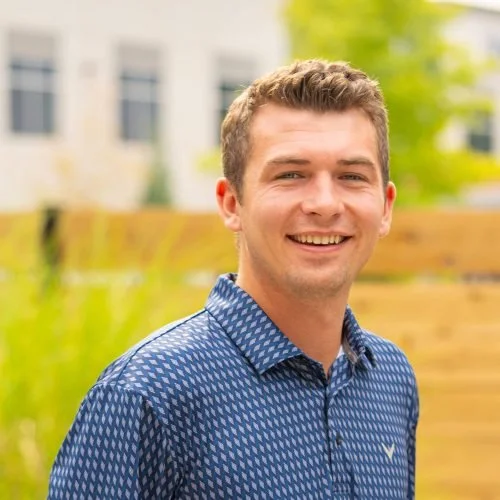 Young man with short brown hair smiling outdoors, wearing a blue patterned button-up shirt, with a blurred background of green foliage and a white building.