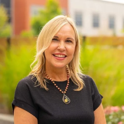 A smiling woman with blonde hair wearing a black top, a beaded necklace, and a pendant, standing outdoors in a park with greenery and blurred buildings in the background.