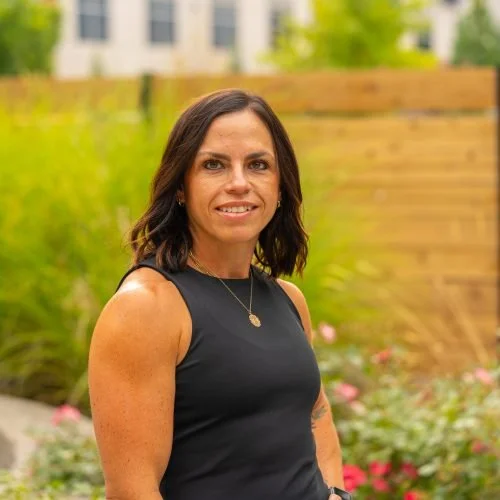 A woman with shoulder-length dark hair wearing a sleeveless black top and a necklace, standing outdoors in a garden area with flowers and a wooden fence in the background.