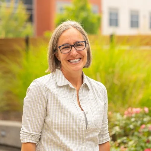 Smiling woman with glasses outdoors in front of greenery and a building.