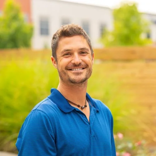 A man smiling outdoors in a blue polo shirt with blurred greenery and a building in the background.