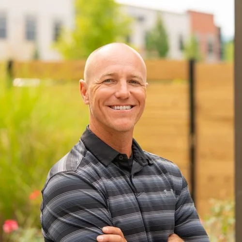 A smiling bald man wearing a black and gray striped polo shirt, standing outdoors in front of a wooden fence and some blurred greenery and buildings in the background.