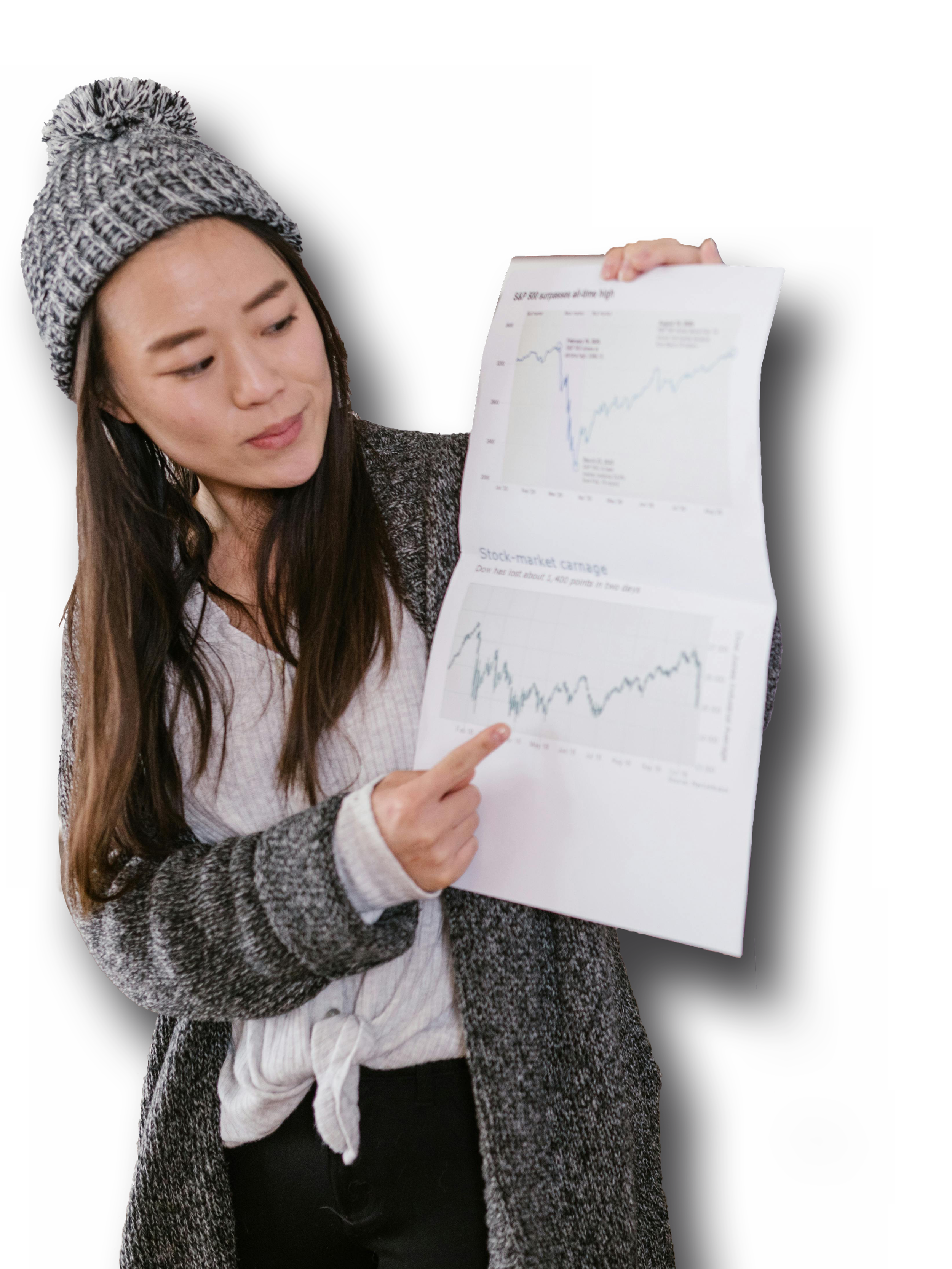 A woman wearing a gray knit hat with a pom-pom and a gray coat is holding and pointing at a large paper with stock market charts and graphs.
