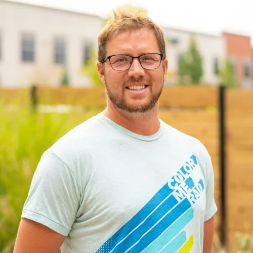 A man with glasses and a beard smiling outdoors, wearing a light blue T-shirt with a colorful design.