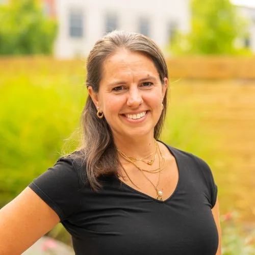 A woman with shoulder-length brown hair smiling outdoors, wearing a black T-shirt and layered gold necklaces, with a blurred background of trees and a building.