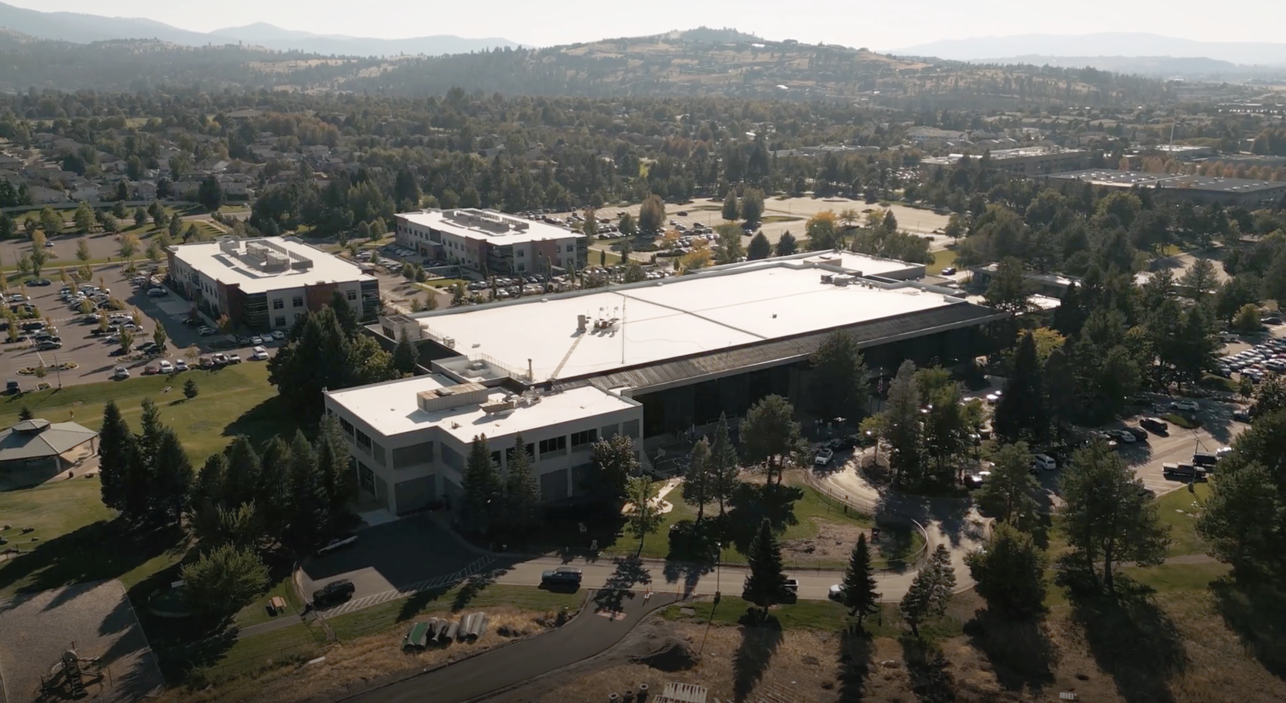 An aerial view of a large building complex with a white roof, surrounded by parking lots and green trees, with a hilly landscape in the background.