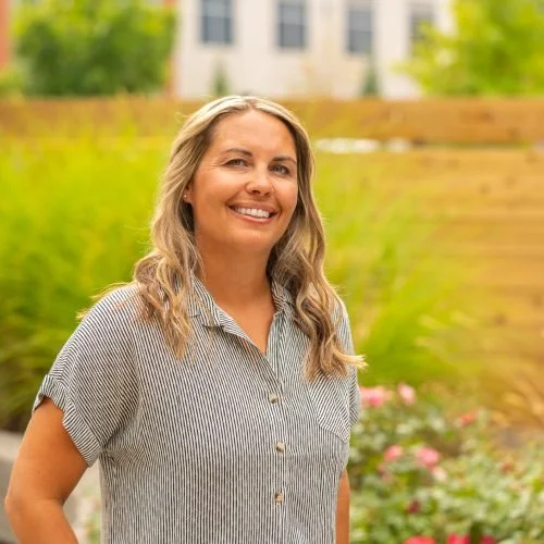 A woman with blonde hair wearing a gray striped shirt, smiling outdoors with a garden and a building in the background.