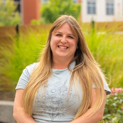 Smiling woman with long blonde hair standing outdoors in front of greenery and a building.