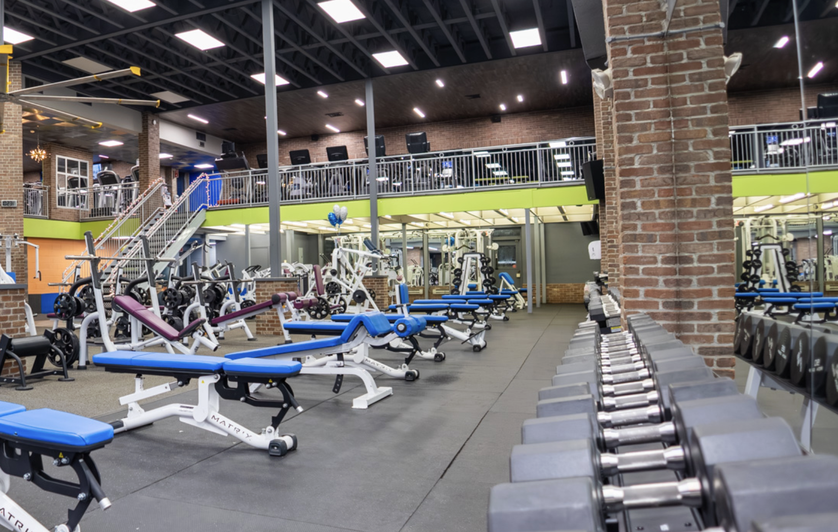 Interior of a modern gym with weightlifting equipment and cardio machines, featuring exposed brick walls, a mezzanine level with additional exercise areas, and balloons near the center.