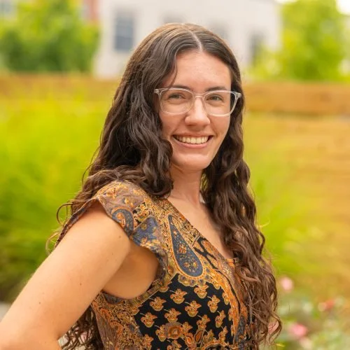 A young woman with long, curly brown hair wearing glasses and a patterned top, smiling outdoors with a blurred green background.