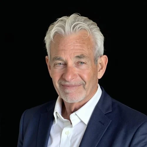 Portrait of an older man with gray hair, wearing a navy blazer and white shirt, smiling against a black background.
