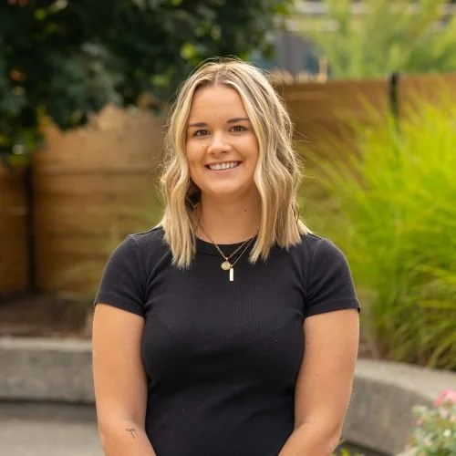 A young woman with shoulder-length blonde hair smiling outdoors in front of a wooden fence and green foliage.