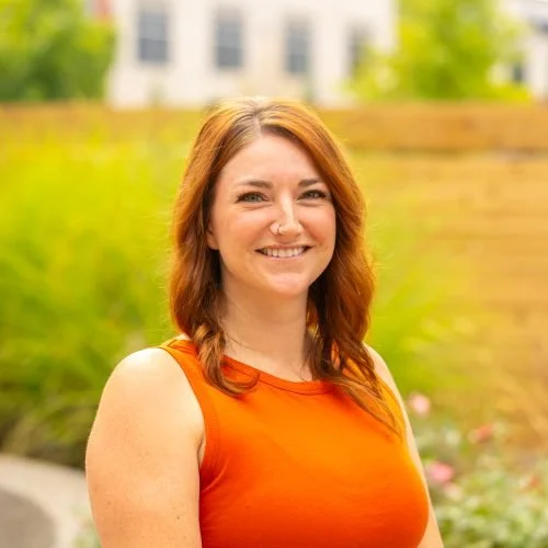 A smiling woman with shoulder-length red hair, wearing an orange sleeveless top outdoors with greenery and a building in the background.