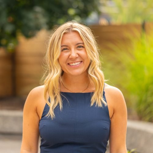 A young woman with blonde hair smiling outdoors, wearing a navy sleeveless top, with a blurred wooden fence and greenery in the background.