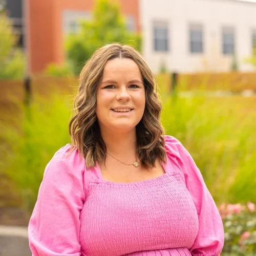 A woman with shoulder-length curly hair smiling outdoors, wearing a pink blouse, with a blurred background of greenery and buildings.