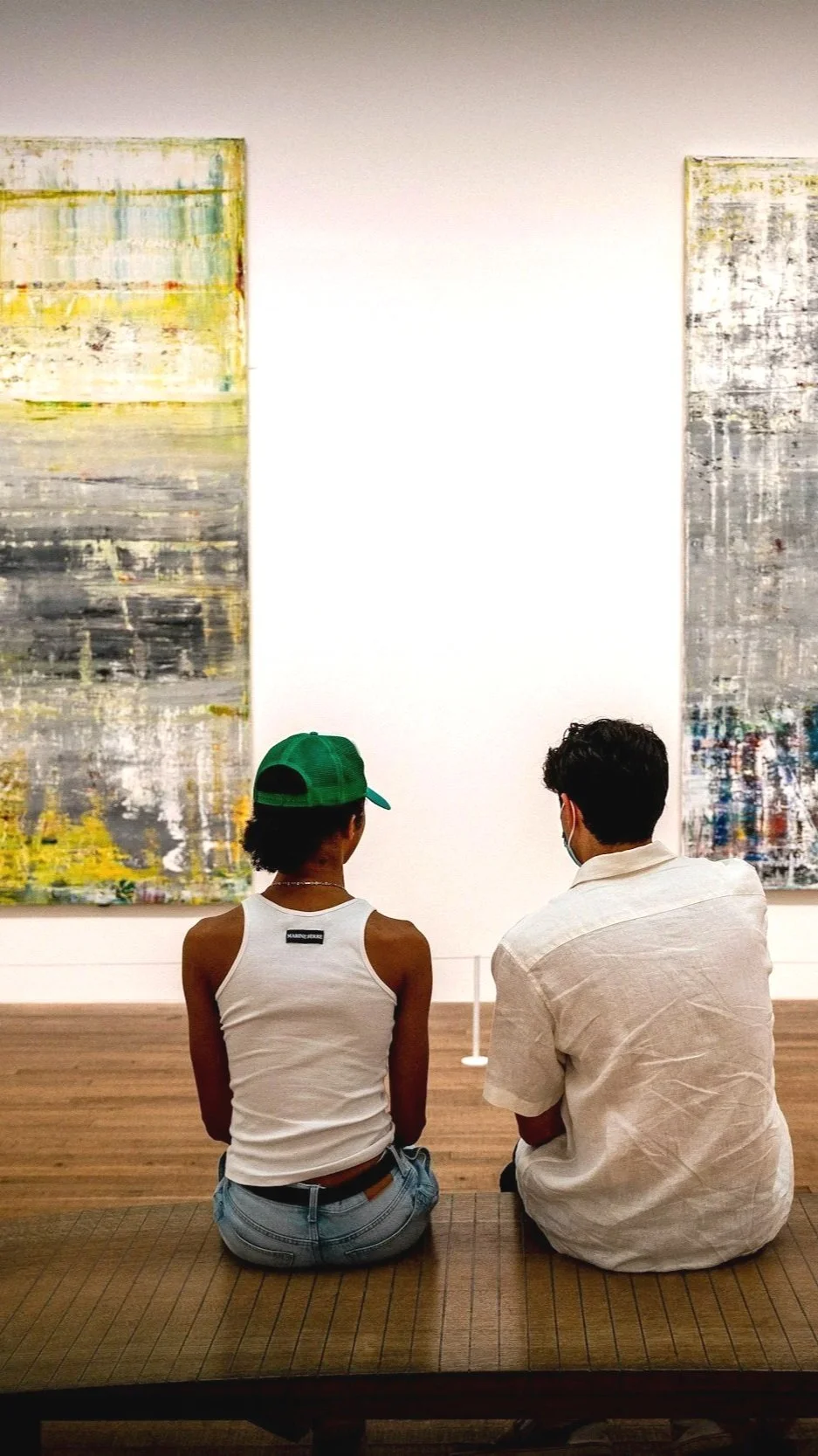 2 people, with their backs to the camera, sit on a bench across from 2 large abstract paintings hanging on a white wall in a gallery.