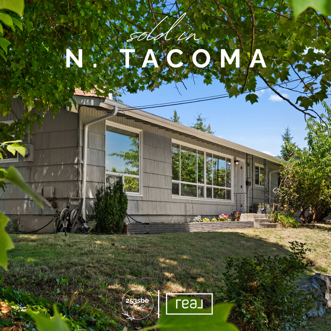 Front view of a single-story house in Tacoma, Washington, with large windows, a small porch with steps, surrounded by trees and shrubs, under a clear blue sky.