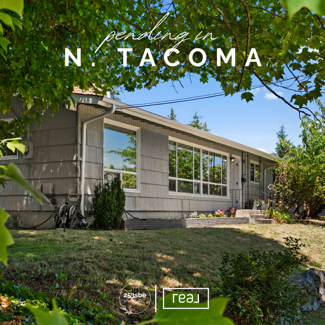 A house with large windows and grey siding, surrounded by lush green trees and plants, with a well-maintained lawn and flowers near the steps leading to the entrance, under a clear blue sky.